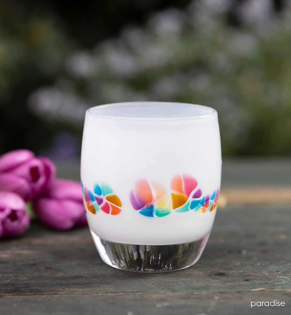 paradise, white rainbow murrini detail hand-blown glass votive candle holder on an outdoor table with pink flowers in background.