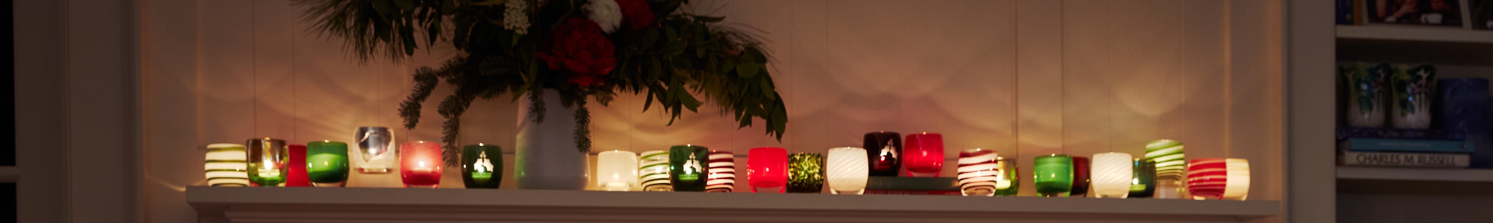 glassybaby holiday collection on a cream mantle with a vase of a floral arrangement.