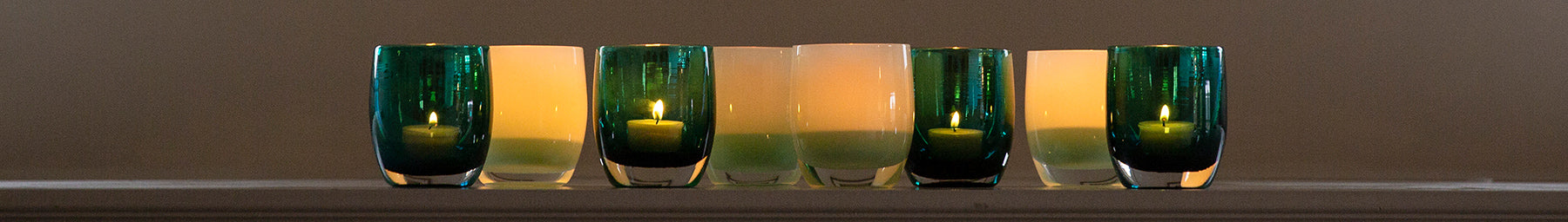 grouping of thank you and gratitude, hand-blown glass votive candle holders with lit tealights sitting on a cream mantle.