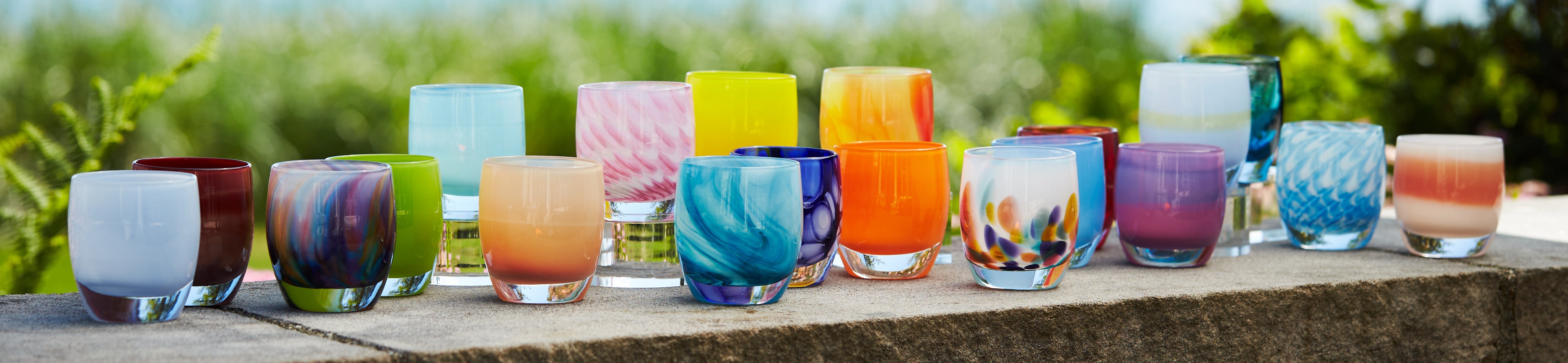 glassybaby summer collection with varying different hand-blown glass votive candle holders sitting on concrete wall with plants and water in background.