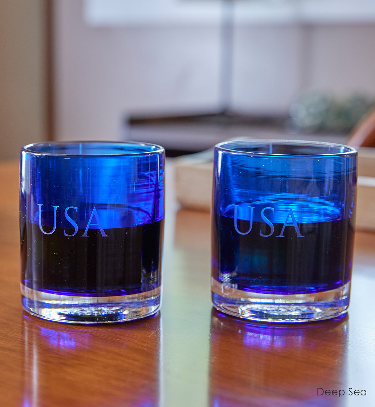 two Deep Sea rockers, royal transparent blue hand-blown  lowball glasses with custom etched "USA" on side. sitting on wood table.
