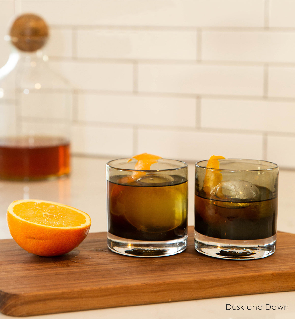 two Dusk and Dawn rockers, dark gray hand-blown glass lowball drinking glasses with ice, liquid and orange peel inside. sitting on a wood cutting board next to an orange with white kitchen background.