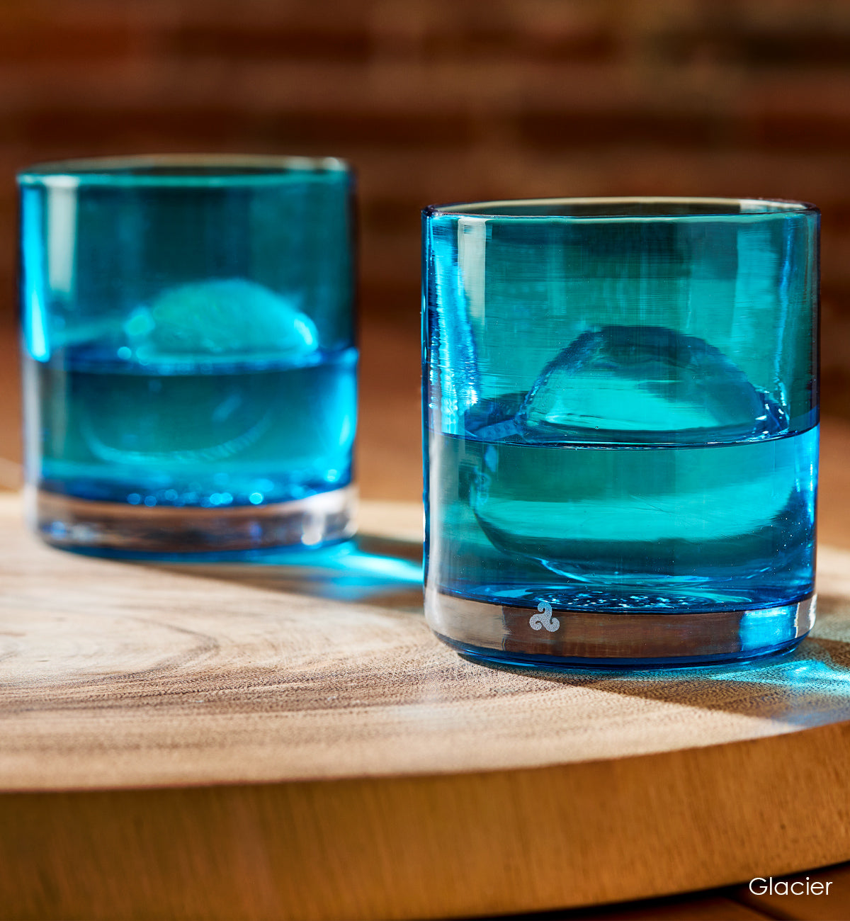 two Glacier rockers, light blue transparent hand-blown glass lowball drinking glasses with liquid and ice atop a light wood table.