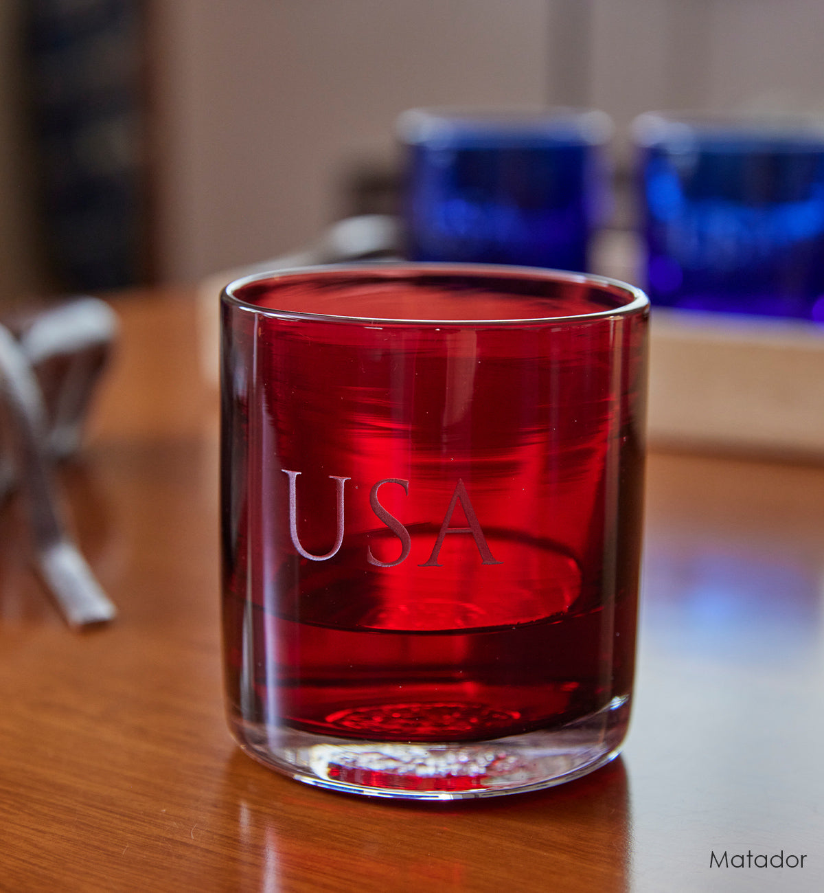 Matador rocker, dark red crimson transparent hand-blown glass lowball drinking glass with the word 'USA' custom etched. sitting on a wood table with two Deep Sea rockers in background.