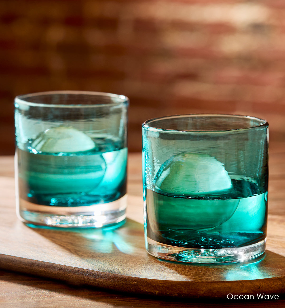 two Ocean Wave rockers, teal blue transparent hand-blown lowball drinking glasses with liquid and ice on top of a wood table and brick wall background.