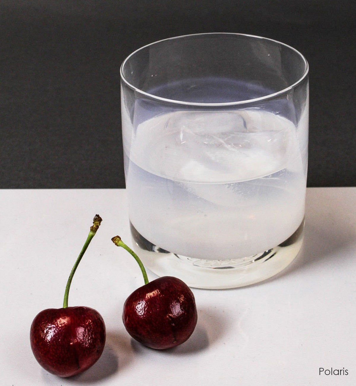 Polaris rocker, white hand-blown glass lowball drinking glass with liquid and ice. sitting atop a white surface next to two cherries.