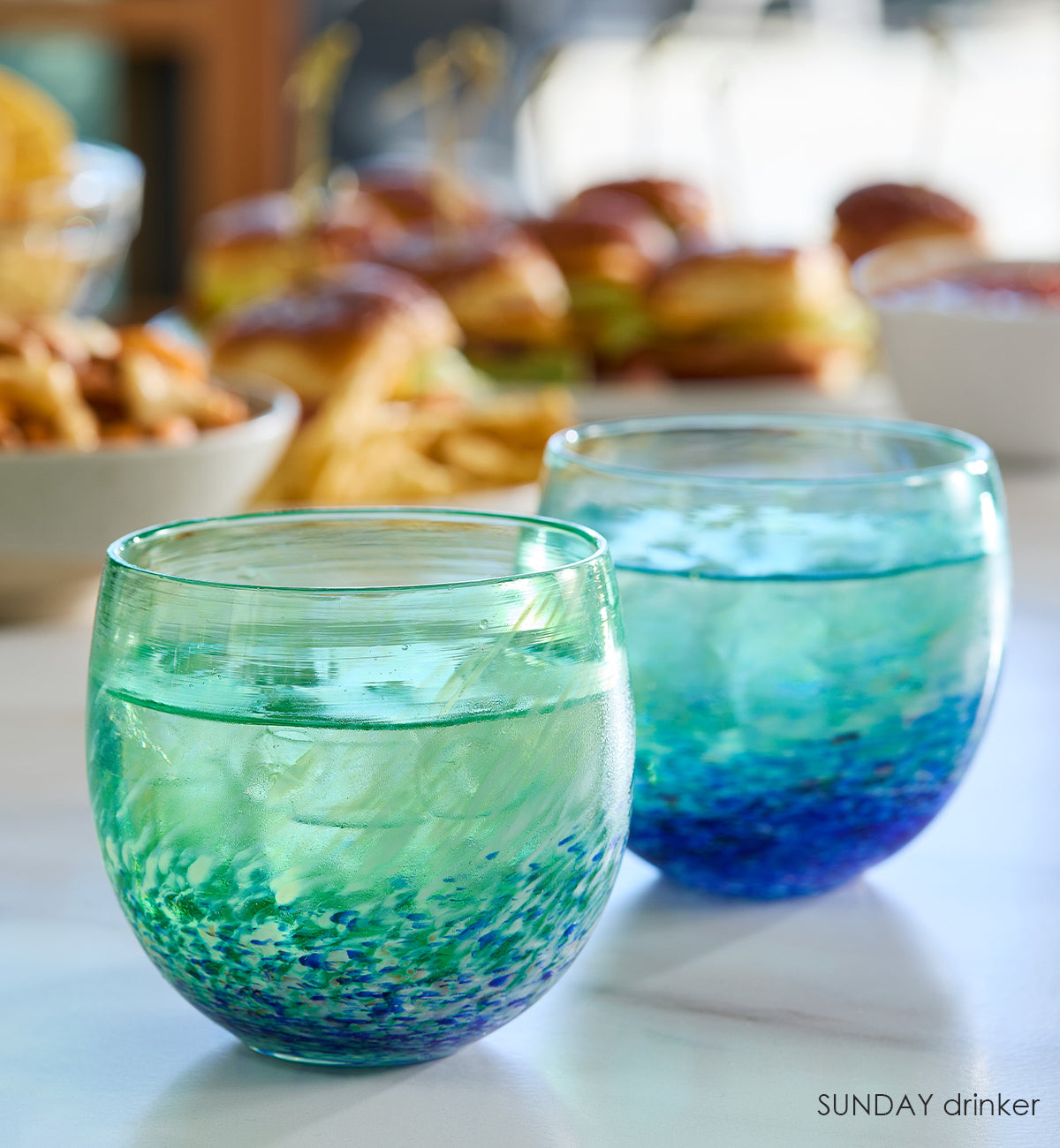 two SUNDAY drinkers, blue green and white swirl hand-blown drinking glasses on a marble surface with watch party food in the background.