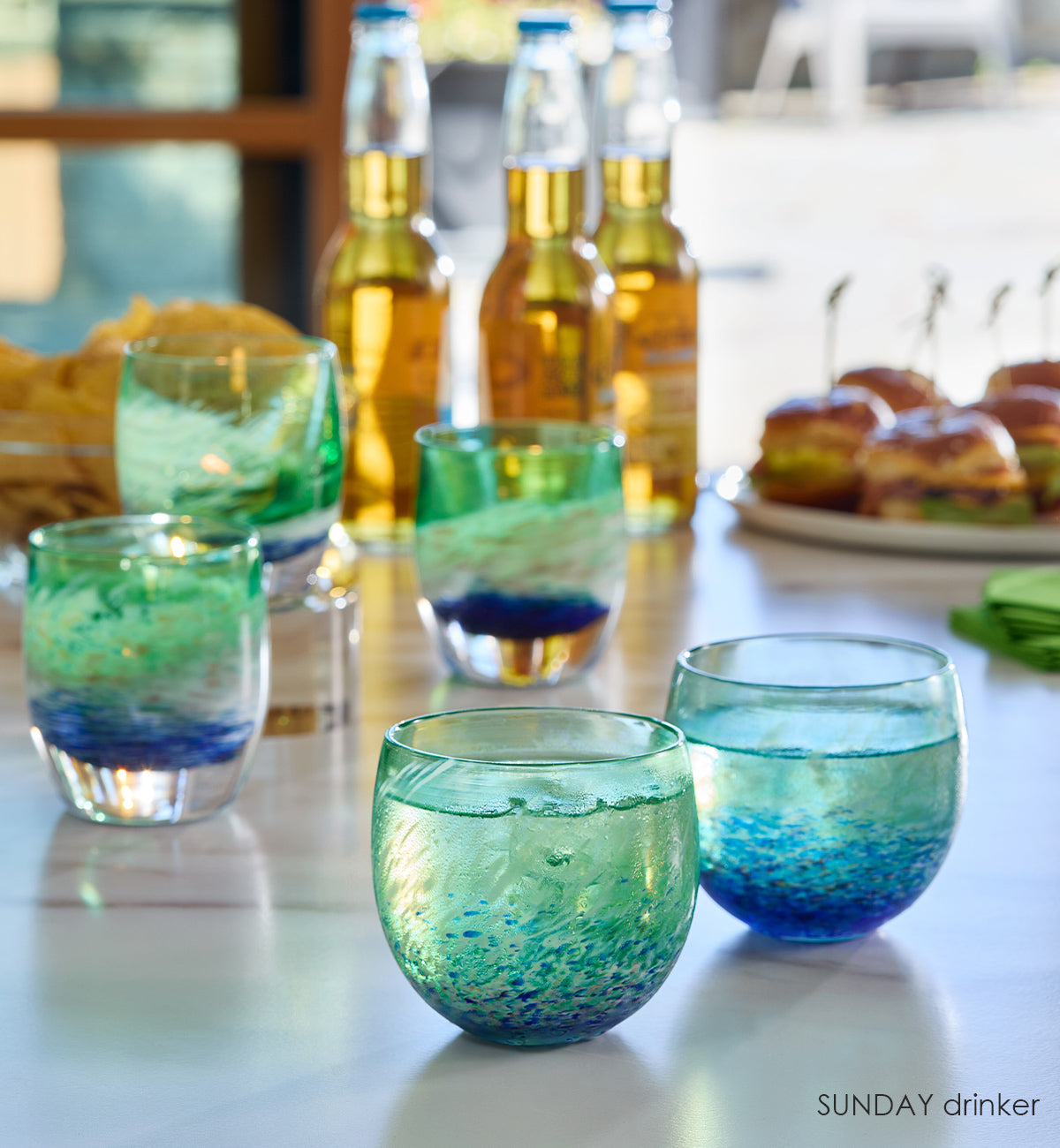 two SUNDAY drinkers, blue green and white swirl hand-blown drinking glasses on a watch party table with SUNDAY votives in the background.