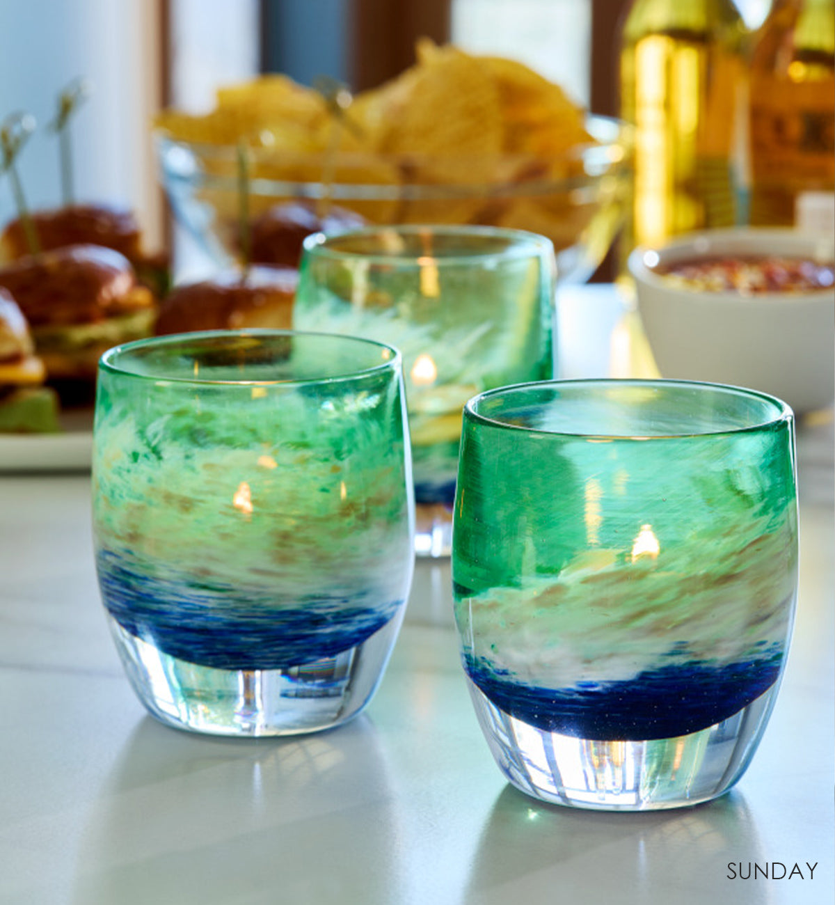 three SUNDAY, blue green and white swirl hand-blown glass votive candle holders on a watch party table with an assortment of snacks and drink.