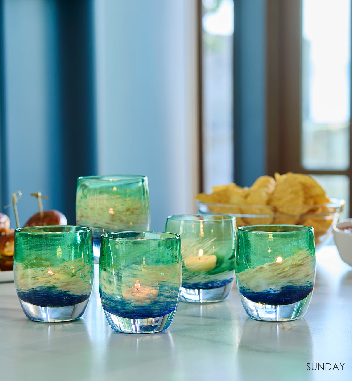 five SUNDAY, blue green and white swirl hand-blown glass votive candle holders on a marble counter with chips and sliders in the background.