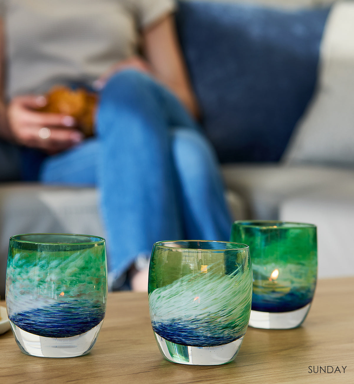 three SUNDAY, blue green and white swirl hand-blown glass votive candle holders on a wood coffee table with two women with drinks sitting behind.