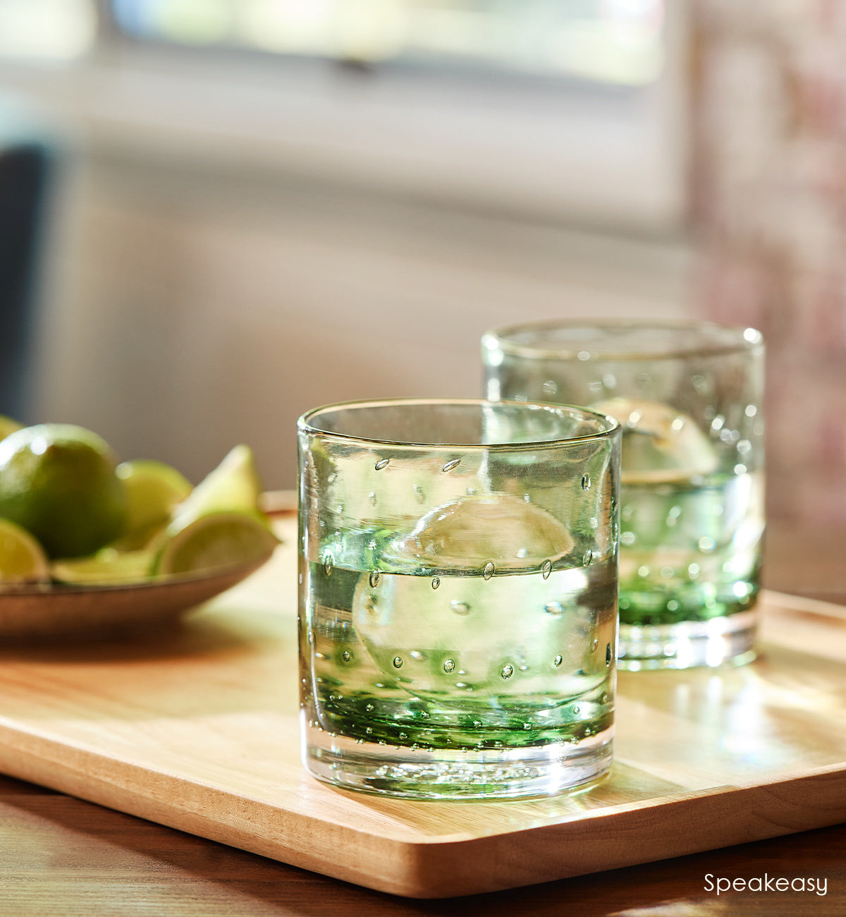 two Speakeasy rockers, dark green hand-blown glass lowball drinking glasses with a bubble pattern. sitting on top of a wood cutting board next to limes.