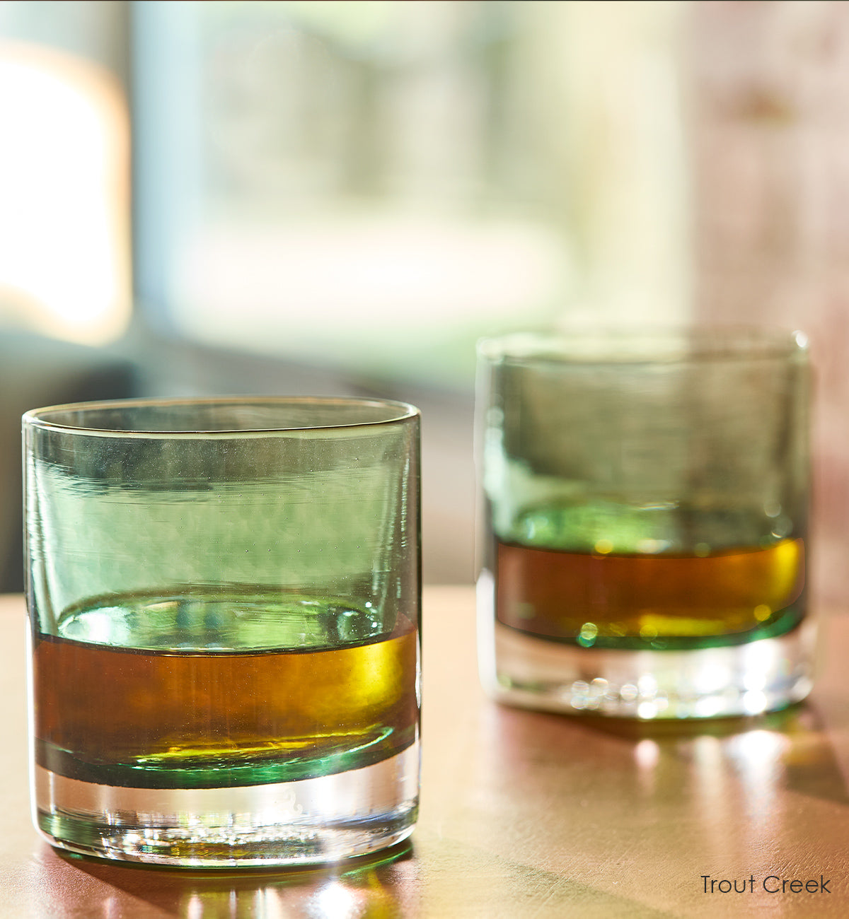 two Trout Creek rockers, dark green teal transparent hand-blown glass lowball drinking glasses with brown liquid inside on a light wood table.