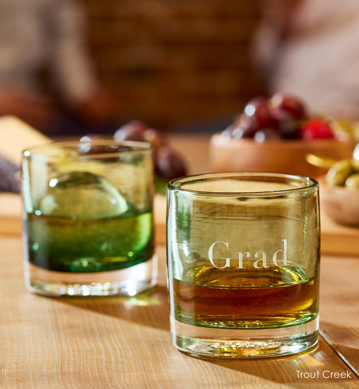 two Trout Creek rockers, dark green teal transparent hand-blown glass lowball drinking glasses, one with "Grad" etched on the side. with ice and liquid inside on a wood table with fruit.