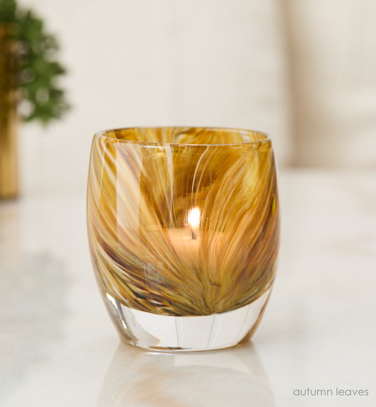 autumn leaves, yellow brown raked hand-blown glass votive candle holder sitting on a reflective marble surface with a plant in the background.