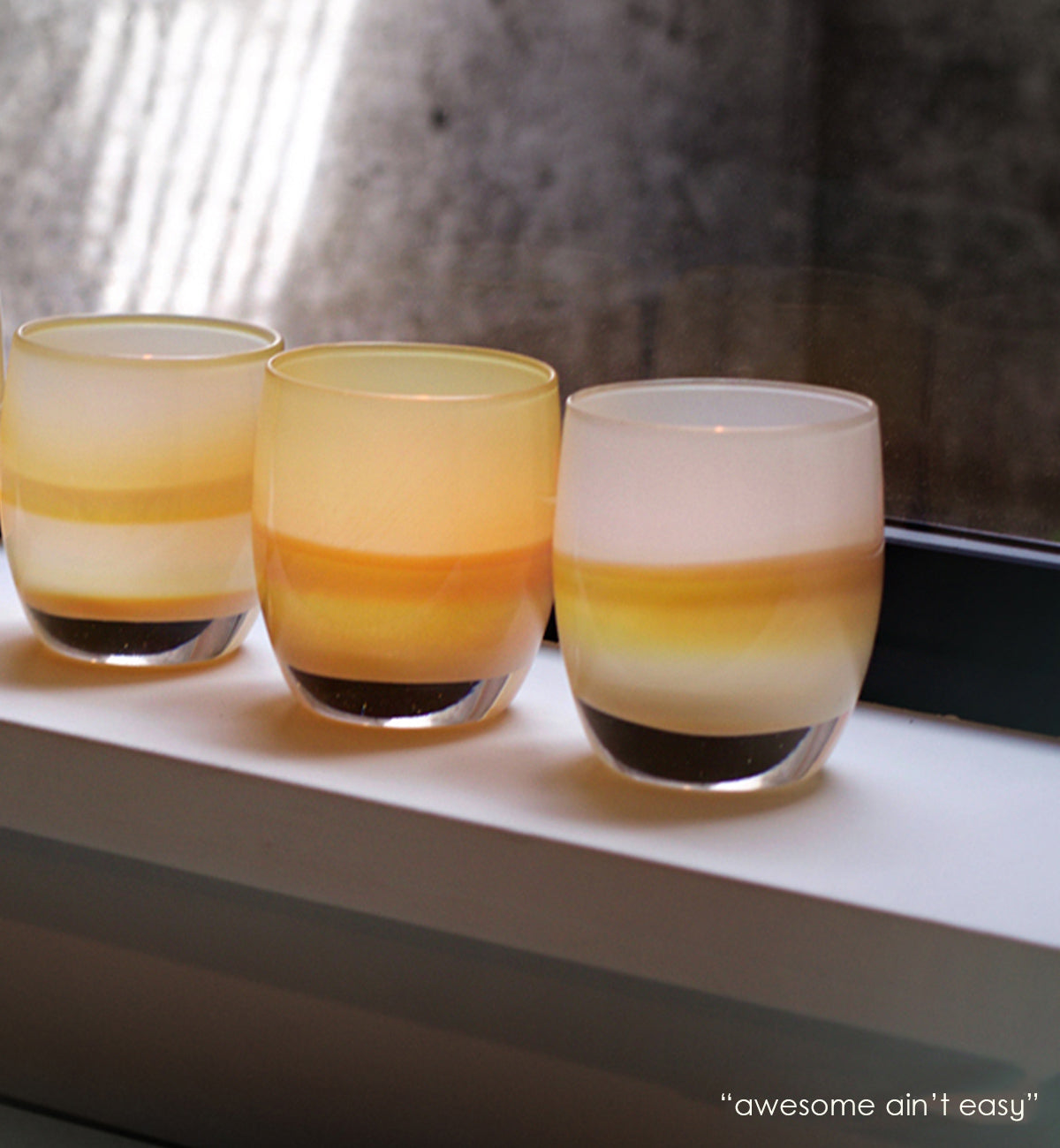 three "awesome ain't easy", sandy yellow hand-blown glass votive candle holders on a white and black windowsill.