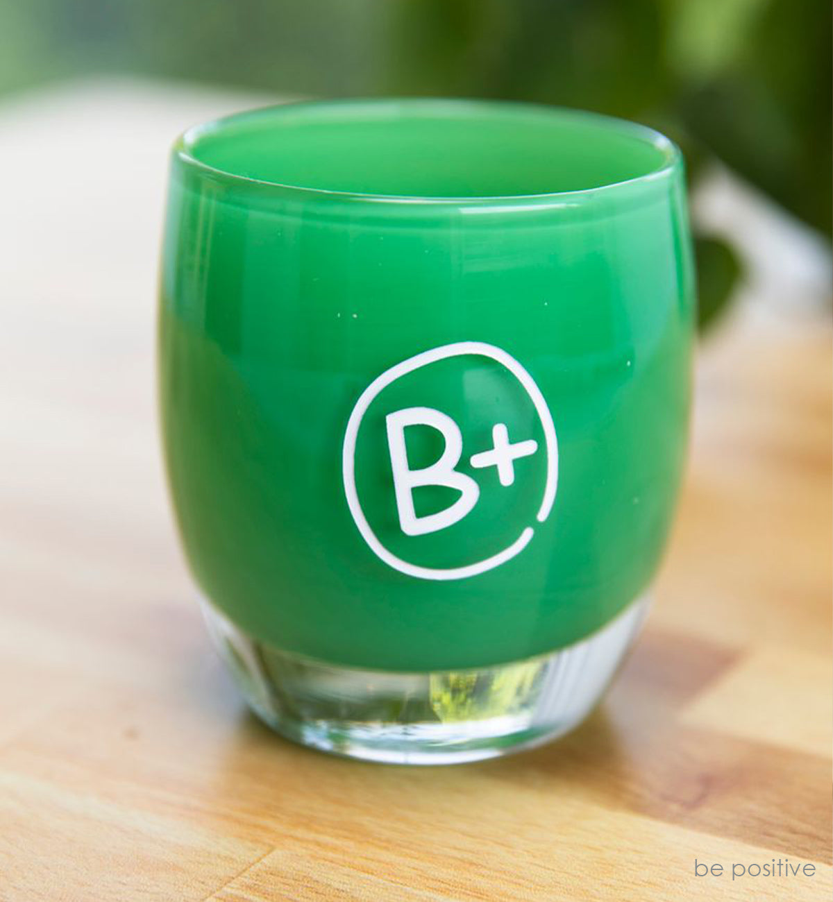 be positive votive, emerald green hand-blown glass votive candle holder with The B+ Foundation logo hand-etched on the side in white paint fill.