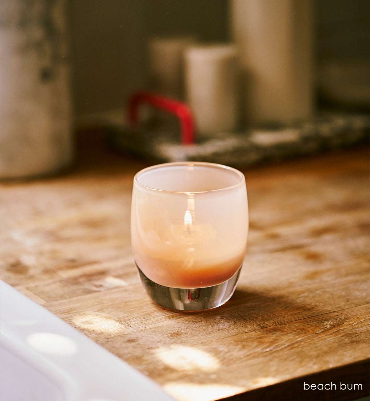 beach bum, tan hand-blown glass votive candle holder on a wood kitchen counter with kitchenware blurred in background.