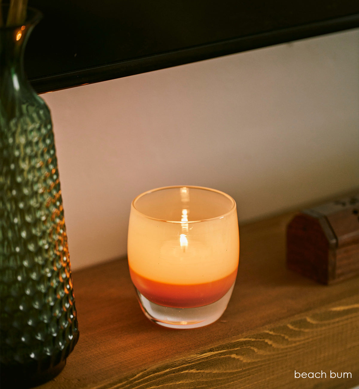 beach bum, tan hand-blown glass votive candle holder on a wood mantle next to a green vase, wood box and screen above.