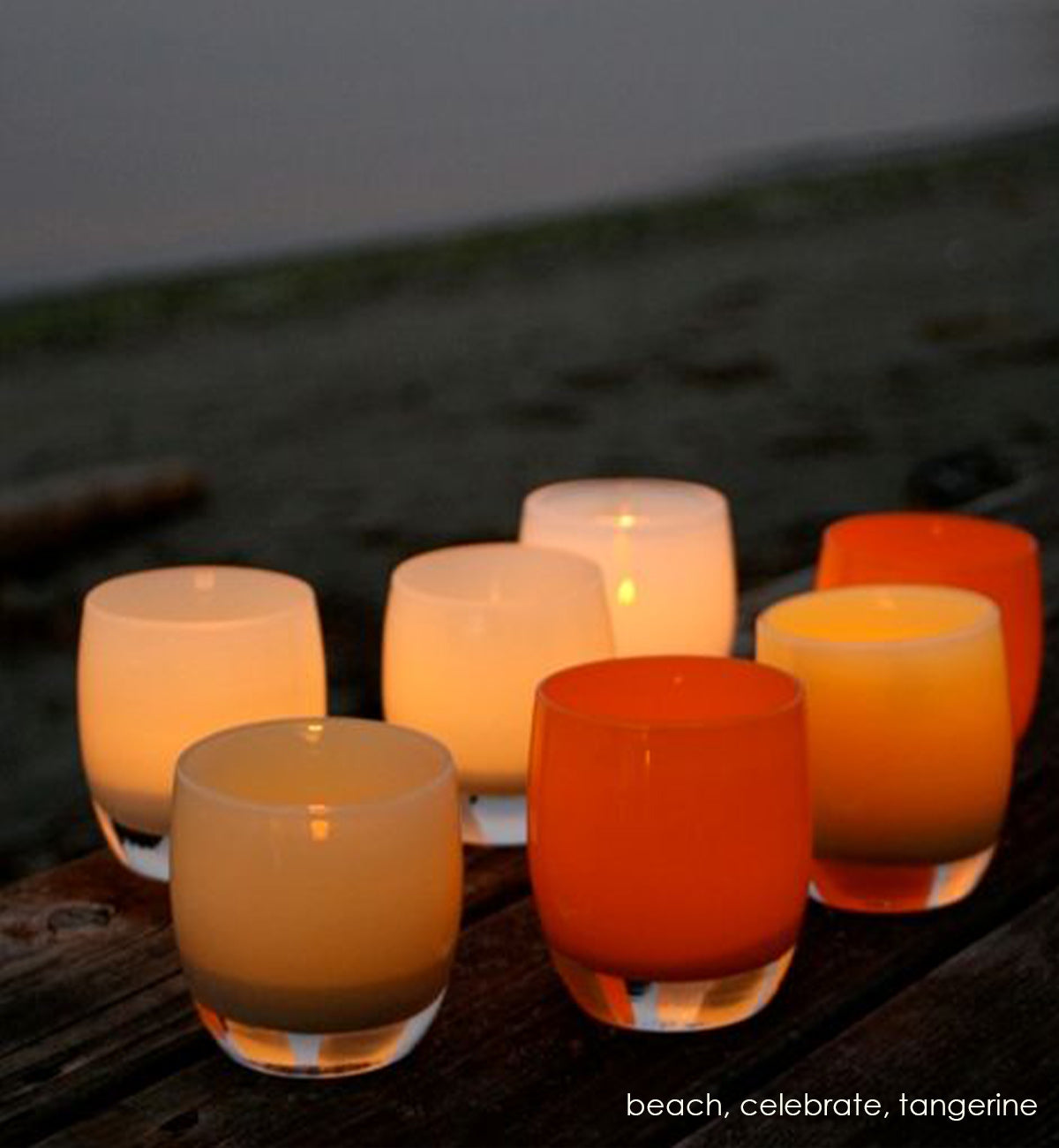 beach sandcastle tan hand-blown glass votive candle holders grouped with tangerine and celebrate on a wood bench on a beach, with the sane and water blurred in background.