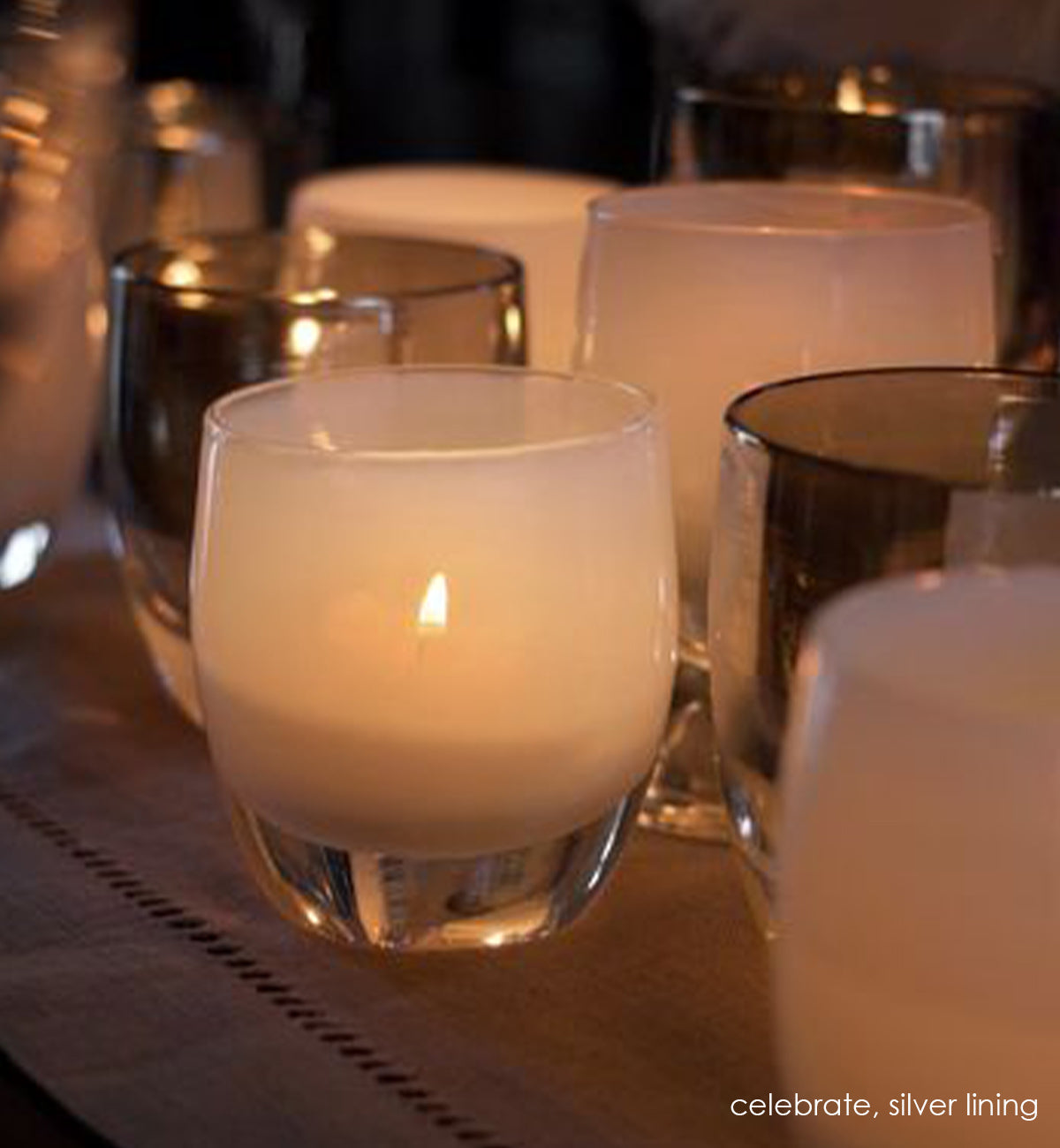 celebrate, snow white hand-blown glass votive candle holder lit with flame on a dining room table runner next to silver lining.