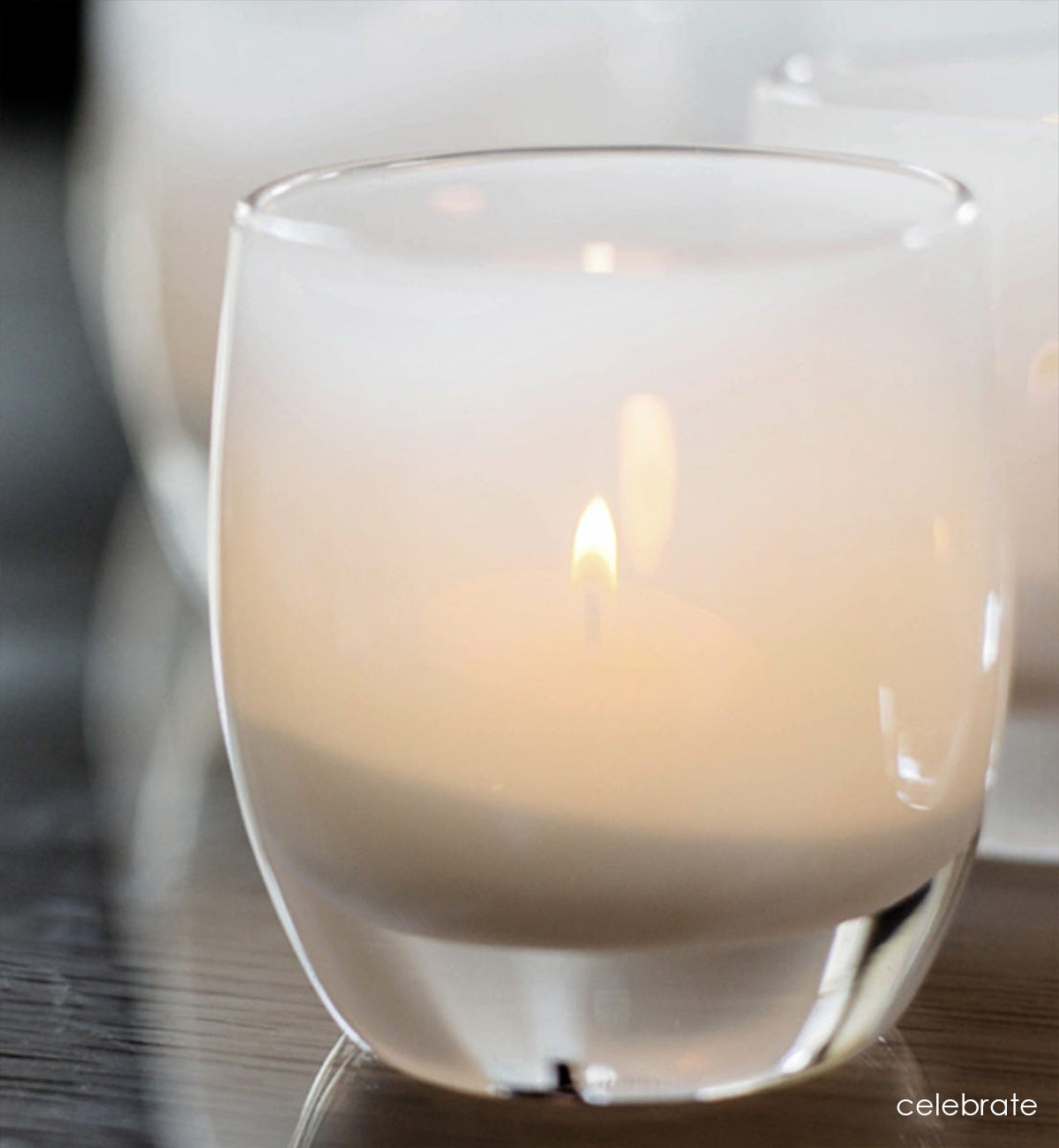 celebrate, snow white hand-blown glass votive candle holder on dark reflective wood table.