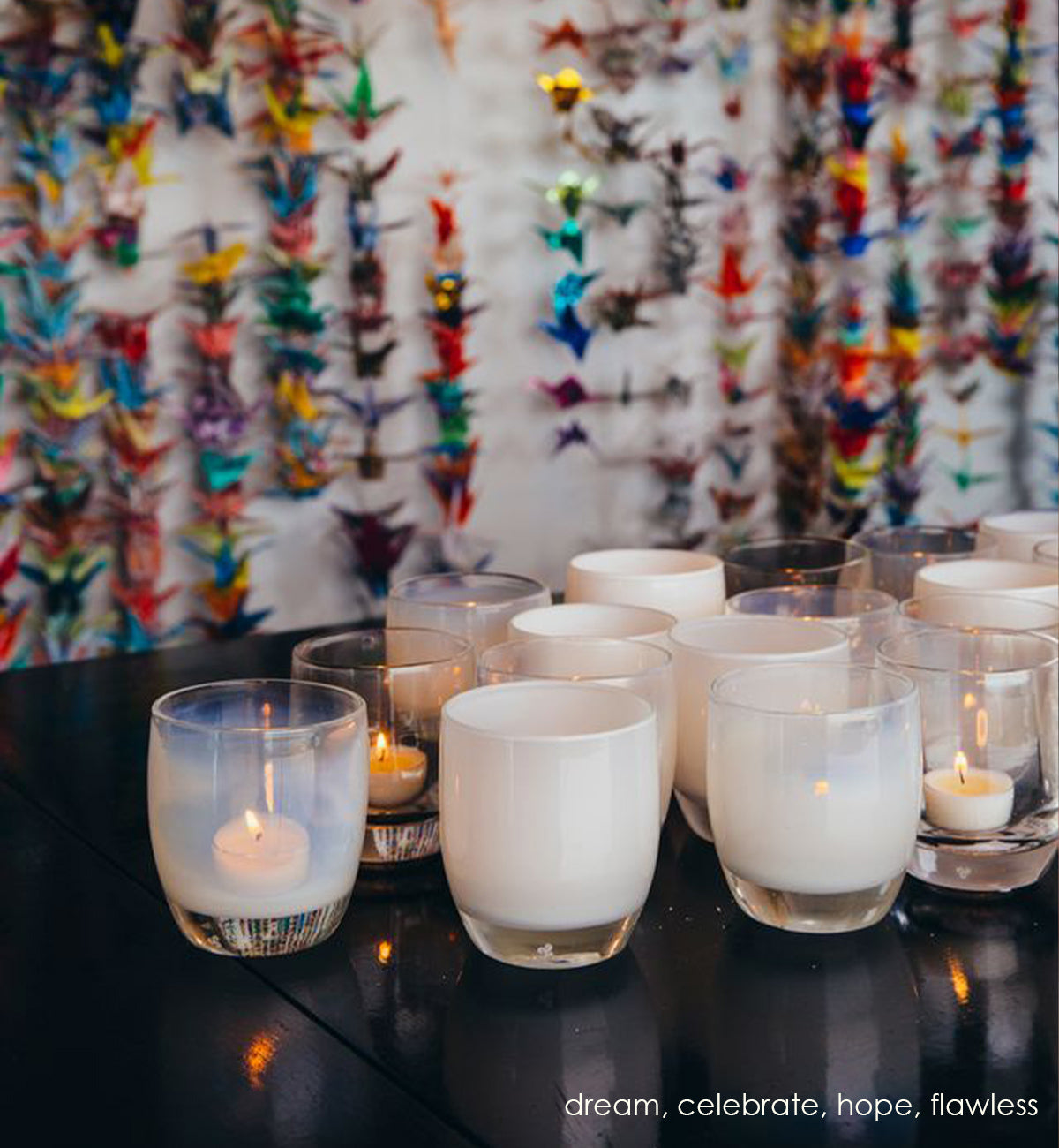 celebrate, snow white hand-blown glass votive candle holder with dream, hope and flawless on a black wood table with hanging paper cranes on background wall.