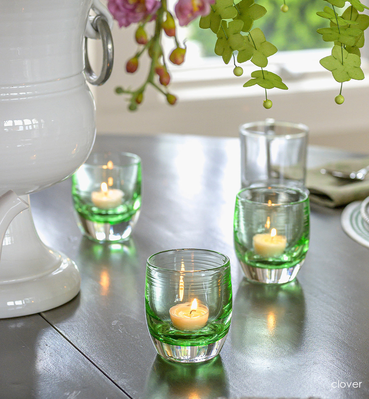 three clover, transparent light green hand-blown glass votive candle holders on a dining room table with a large vase of flowers and diningware.