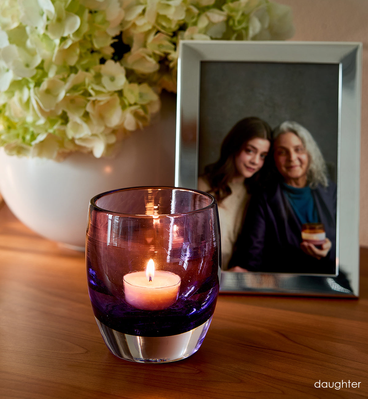 'daughter' lit purple hand-blown glass candle holder on wood dresser. Photo of  mother and daughter in silver photo frame and flowers in white vase in background.