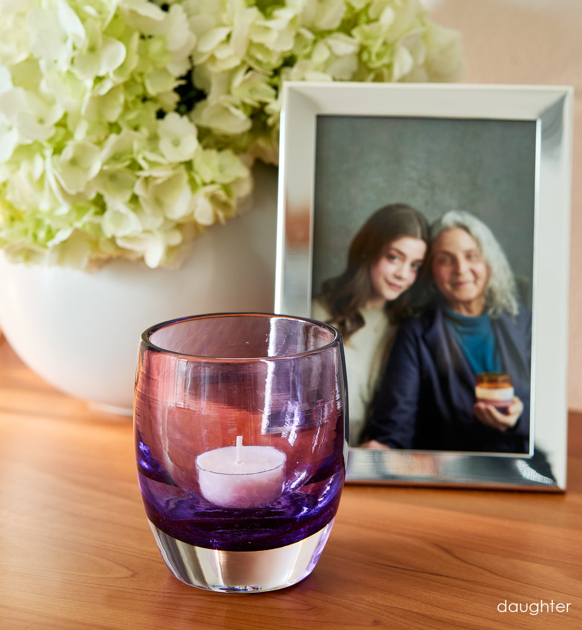 'daughter' purple hand-blown glass candle holder on wood dresser. Photo of  mother and daughter in silver photo frame and flowers in white vase in background.