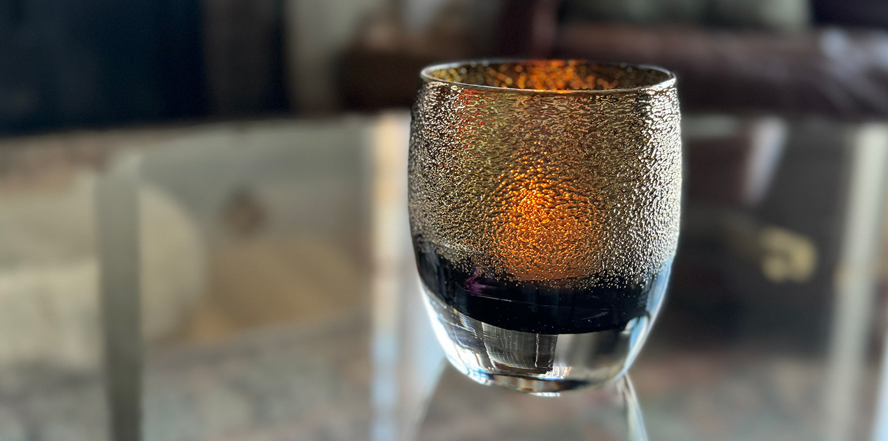 disco, black with gold shimmer handblown glass votive candle holder glowing with a lit tealight, on a clear glass coffee table in a living room.