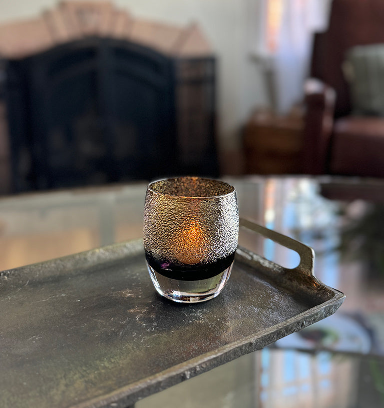 disco ball, black with gold shimmer hand-blown glass votive candle holder glowing with a lit tealight, sitting on a metal tray on a glass coffee table in a living room.