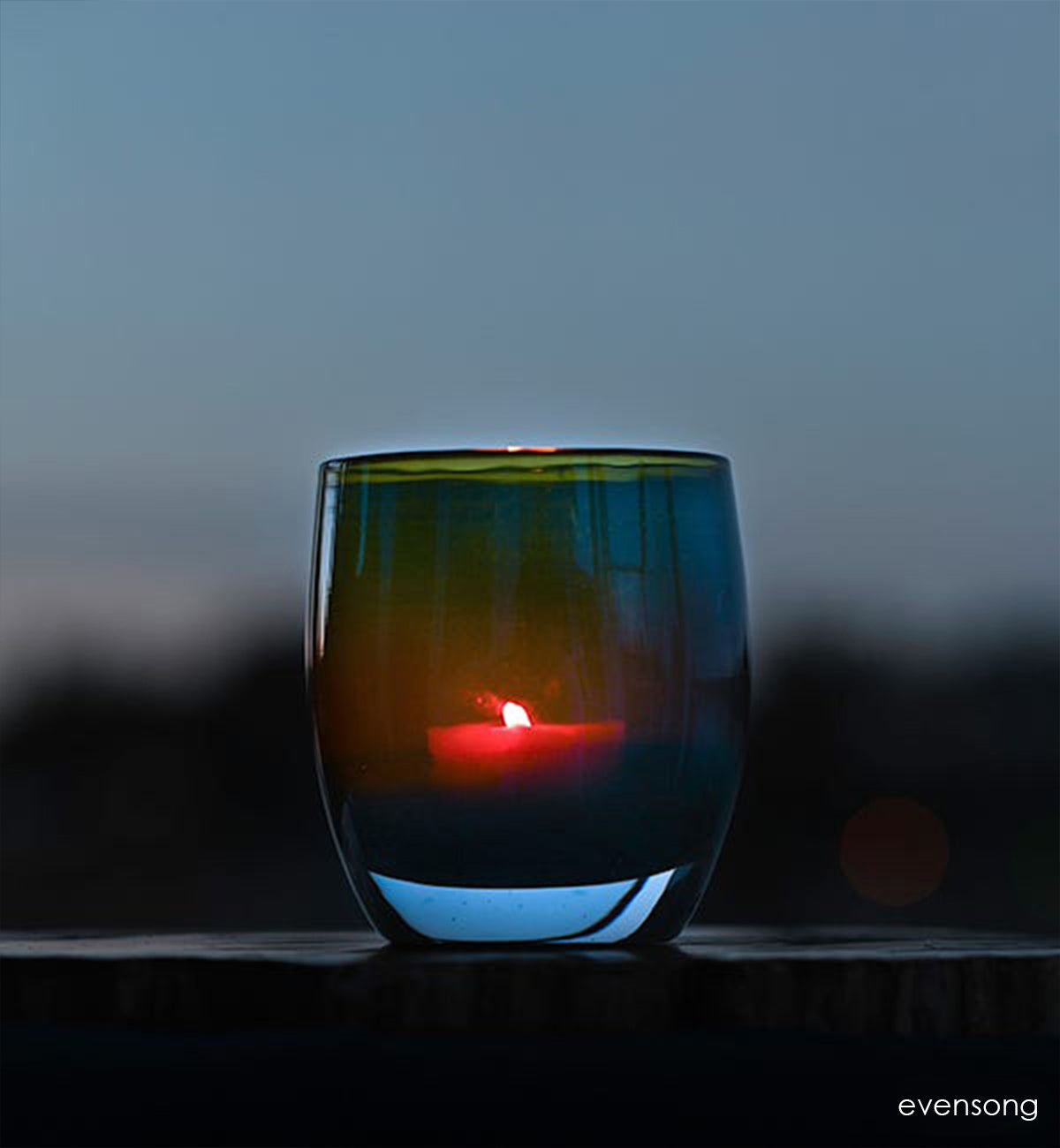 evensong hand-blown green, blue and brown glass votive candle holder lit with a red flame on an outdoor mantle.