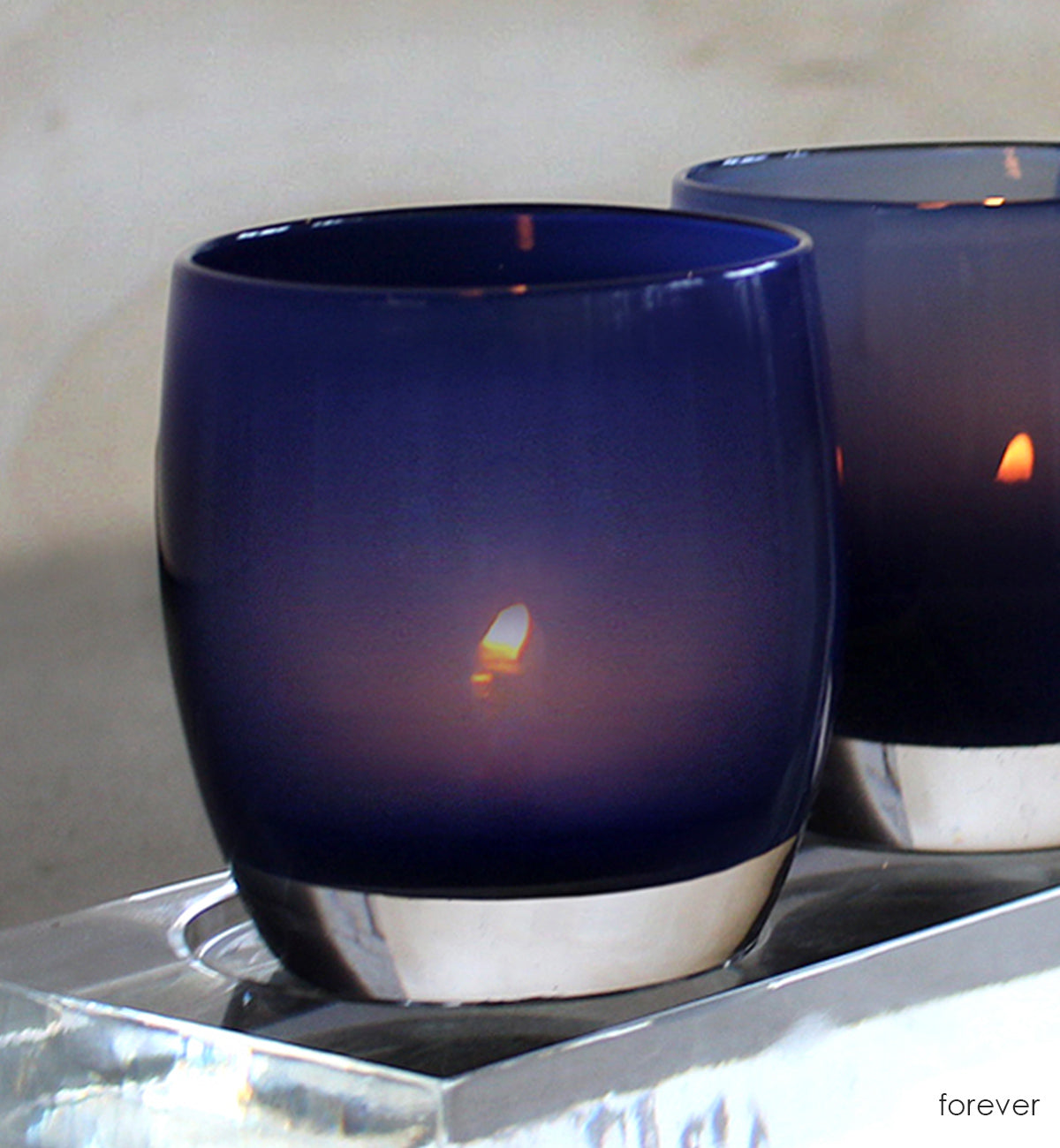 two forever deep navy blue hand-blown glass votive candle holders lit on a square acrylic babystand.