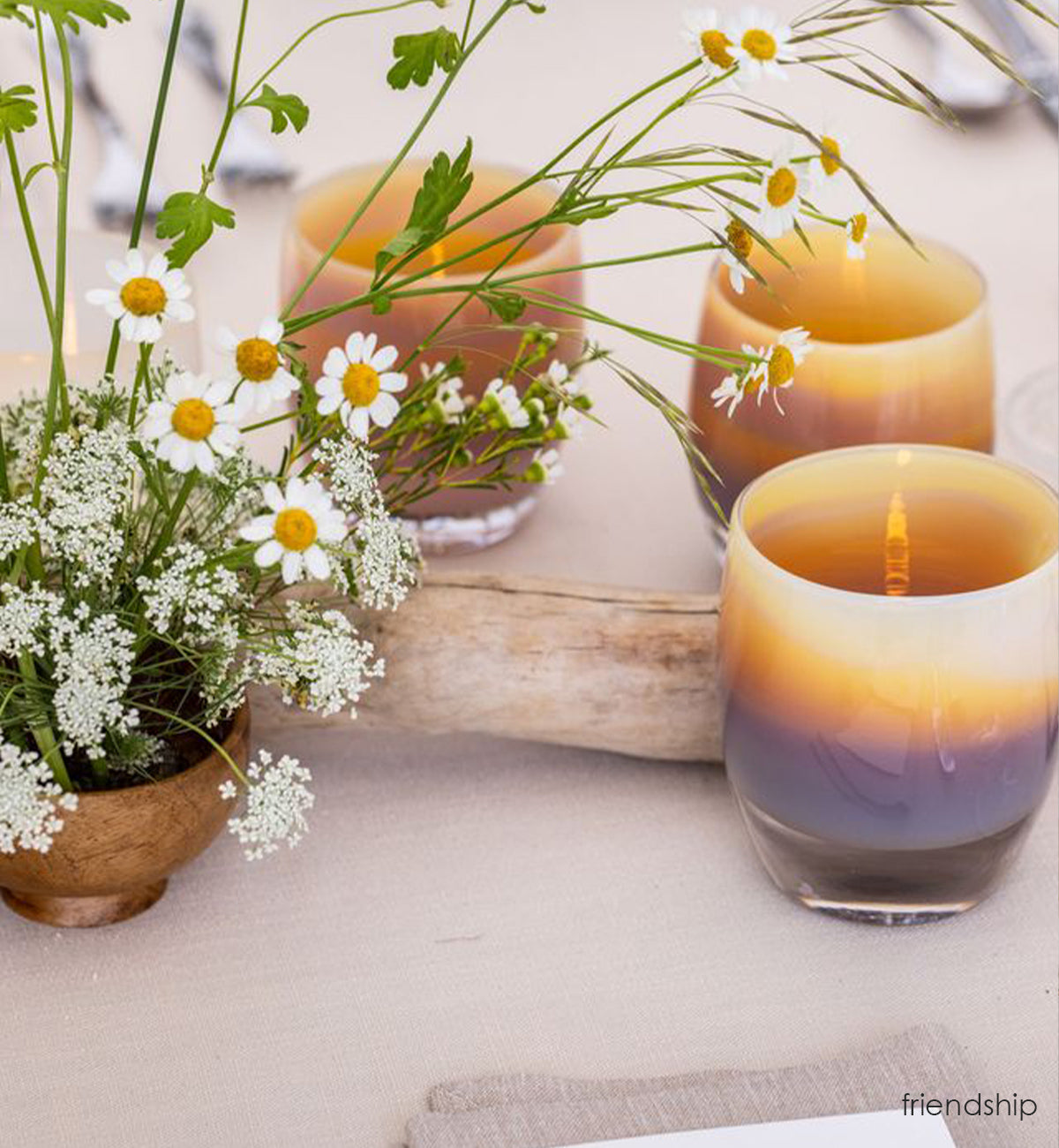 three friendship, hand-blown soft yellow with brown and purple banding glass votive candle holders on a wedding dining table with vased daisies and beach wood.