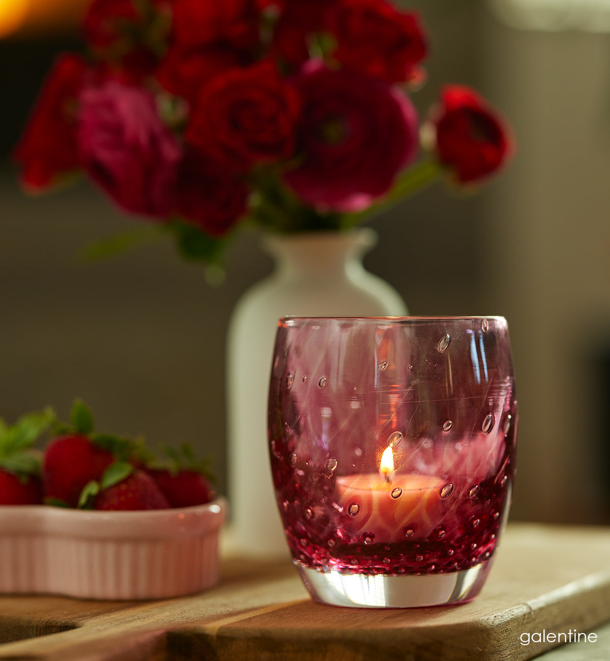 'galentine' is a beautiful hand-blown glass votive with a bubble pattern in red. Sitting on wood cutting board with bowl of strawberries in white dish  and red roses in white vase behind it. 