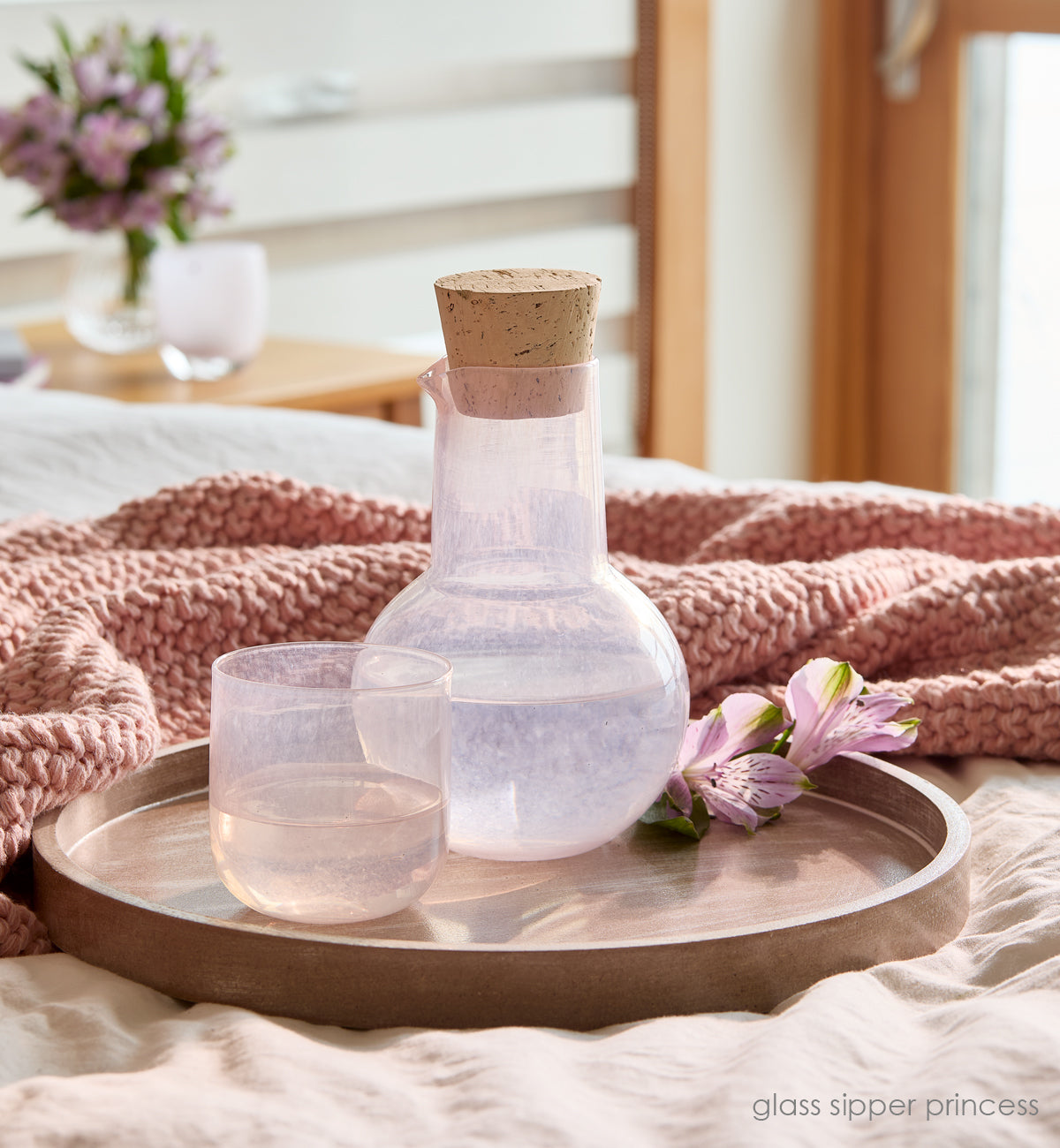 glass sipper princess, light pink hand-blown glass and carafe with a cork on wood tray on bed with pink blanket.