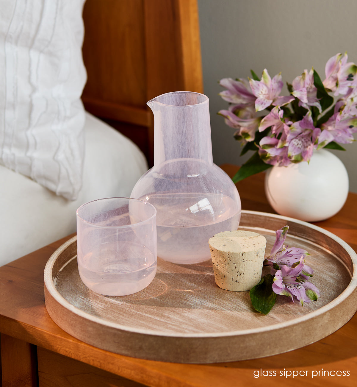 glass sipper princess, light pink hand-blown glass and carafe on wood tray at bedside table with purple flowers.