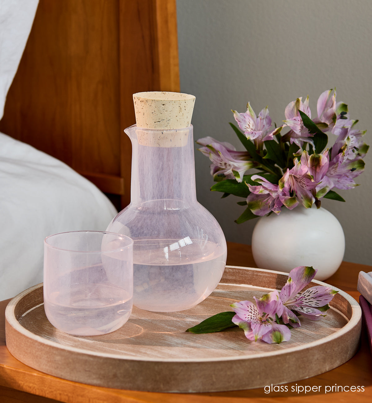 glass sipper princess, light pink bubble, hand-blown glass and carafe with a cork on wood tray at bedside table with purple flowers.