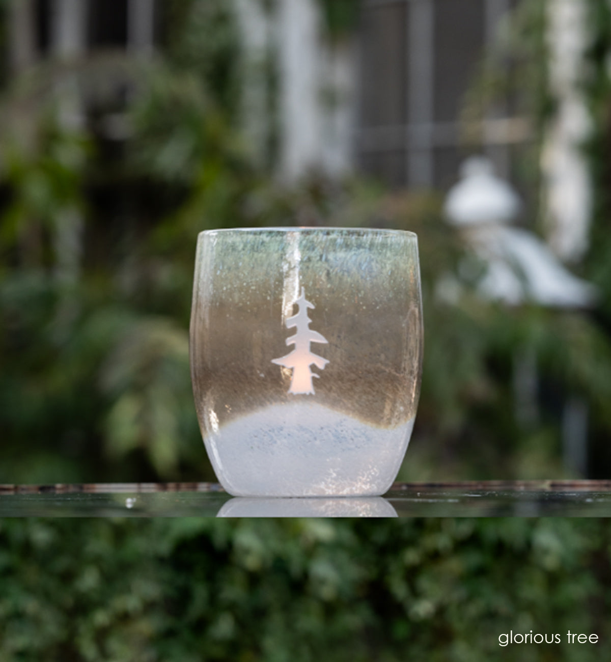 glorious brown white etched tree hand-blown glass votive candle holder sitting on a reflective surface with ivy leaves and trees blurred in the background.