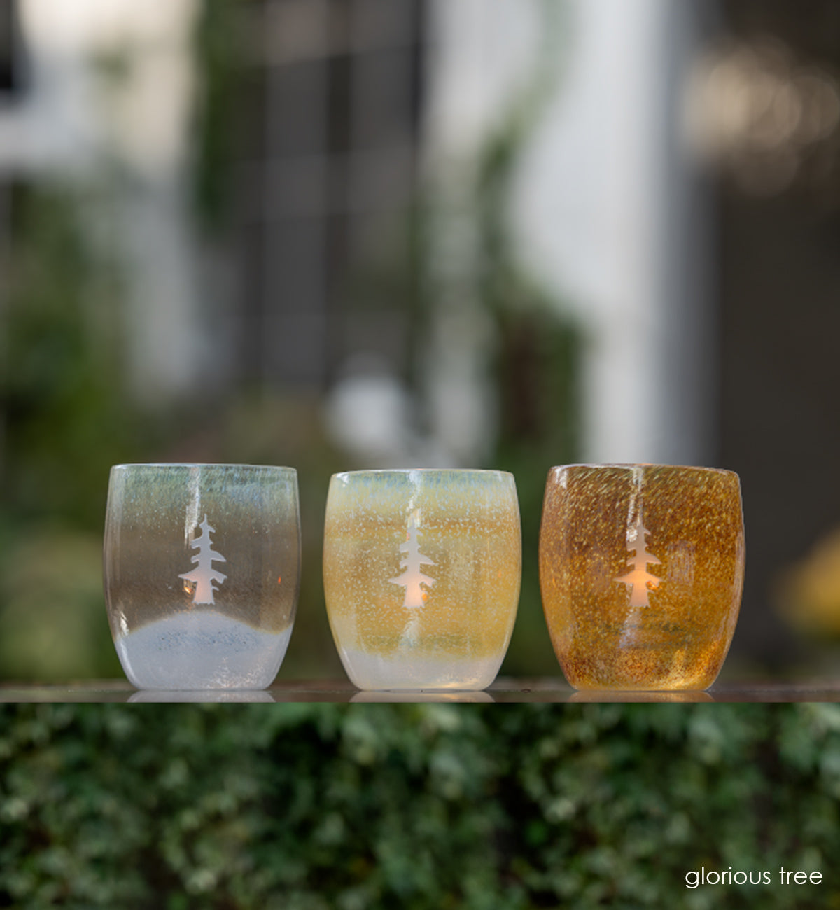 three glorious brown white etched tree hand-blown glass votive candle holders on a clear surface with ivy leaves, trees and a white house blurred in background.