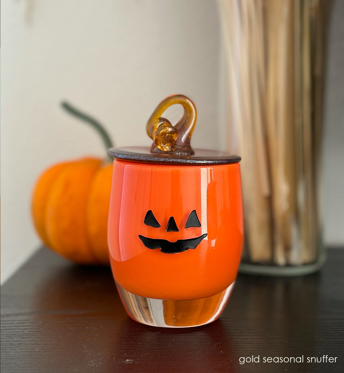 gold seasonal snuffer, gold hand-blown glass votive snuffer in shape of top of a pumpkin and stem sitting on jack-o-lantern on dark wood surface with pumpkin in background.