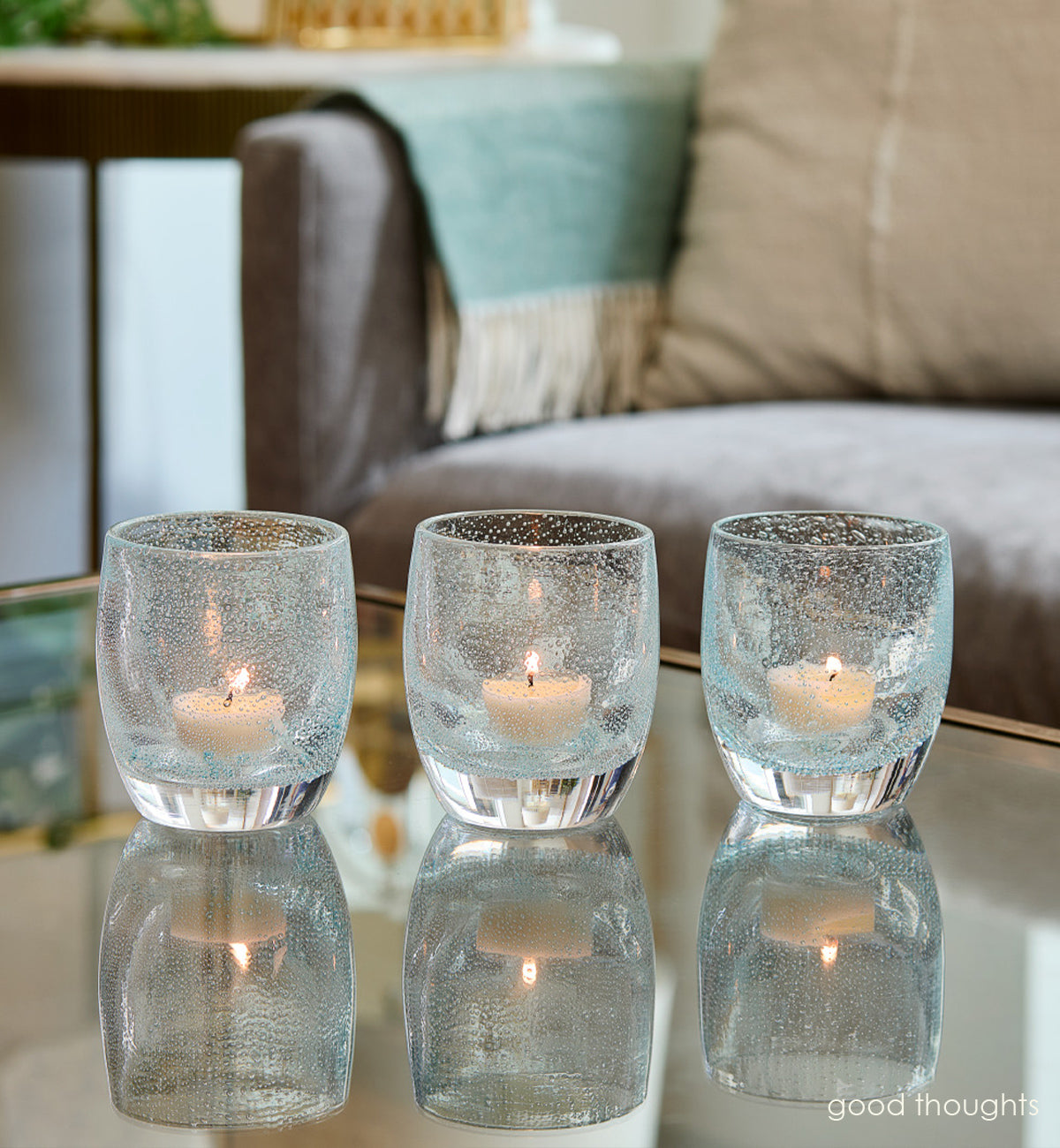 three good thoughts,  transparent light blue with small bubble pattern hand-blown glass votive candle holders on a reflection coffee table with couch and side table in background.