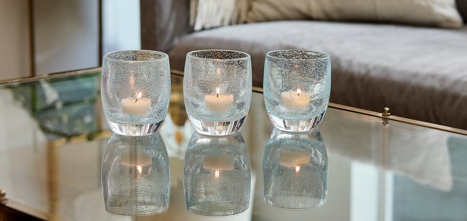 three good thoughts, light clear blue with bubbles hand-blown glass votive candle holder on a reflective coffee table with lit tealights. gray couch in background.