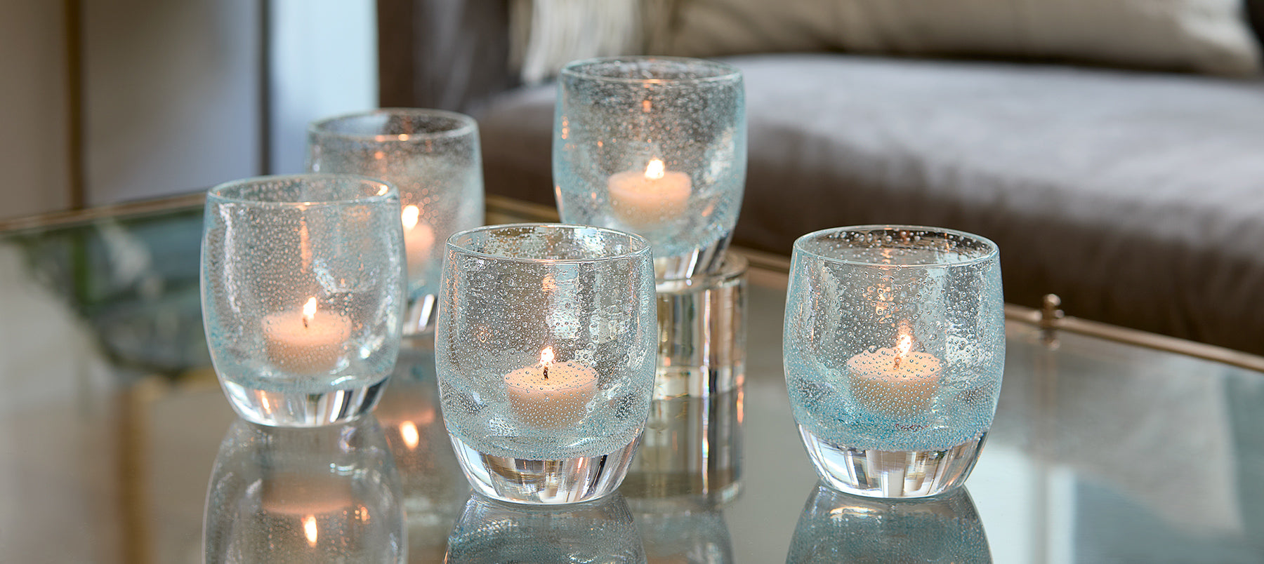 five good thoughts, light clear blue with bubbles hand-blown glass votive candles holders with lit tealights on a reflective coffee table.