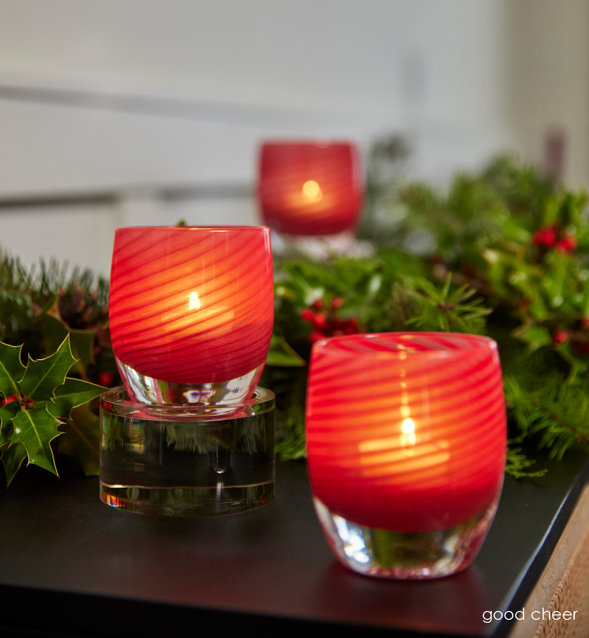 three lit red swirls on translucent hand-blown glass candle holder on wood desk with green holly leaves.