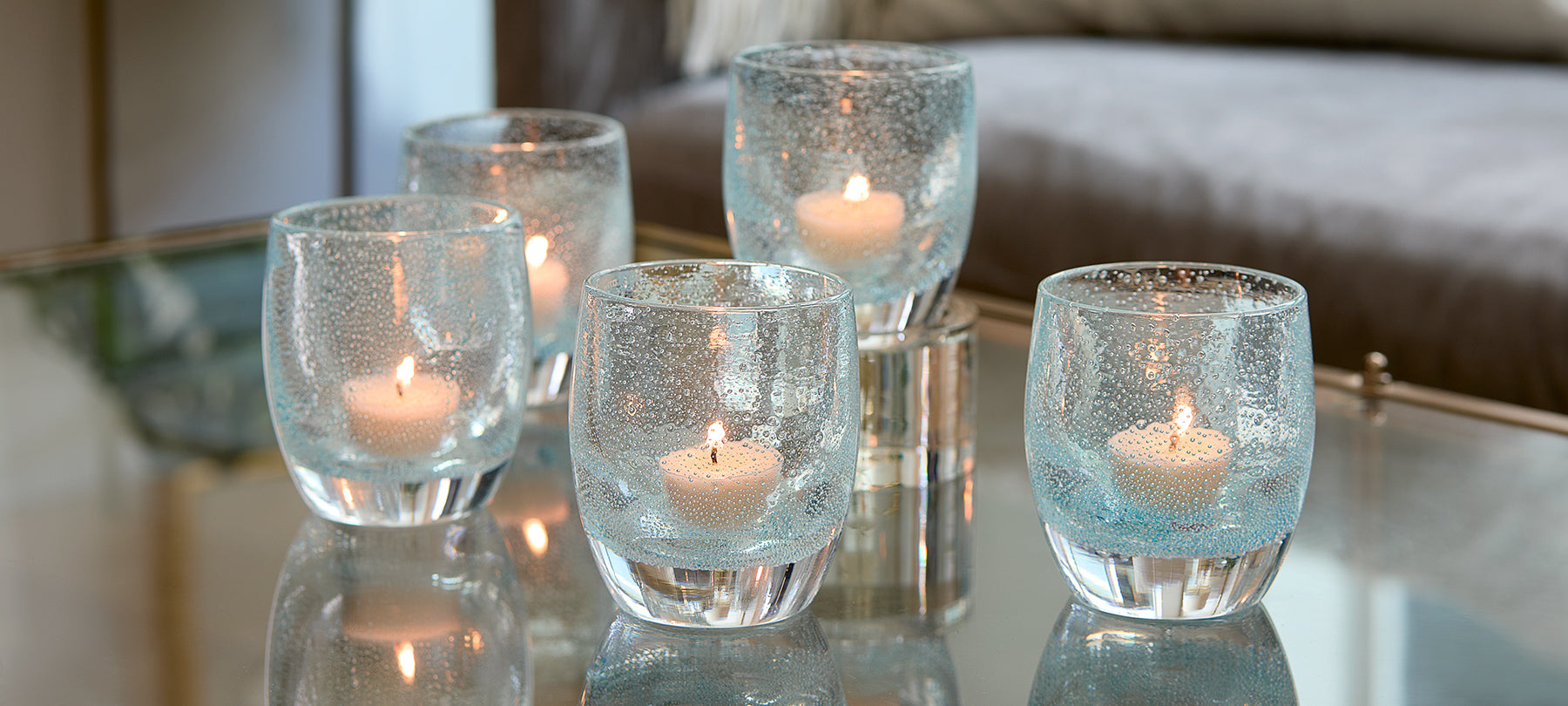 five good thoughts, transparent light blue with small bubble pattern hand-blown glass votive candle holders on a reflective glass coffee table with couch in background.