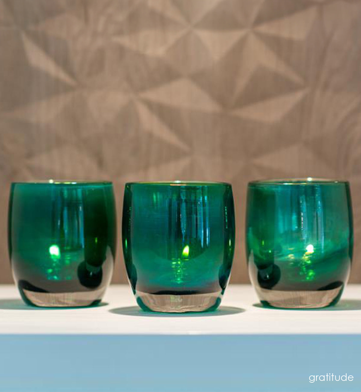  three gratitude, textured translucent teal green with silver luster, hand-blown glass votive candle holder on white shelf.