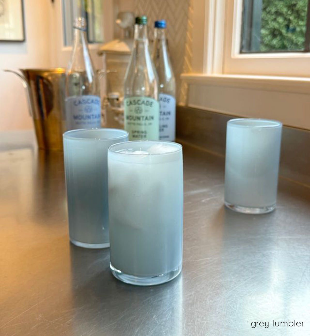 three grey tumblers, opaque light grey hand-blown water glasses with liquid and ice in each on a kitchen counter with sparkling water bottles in background.