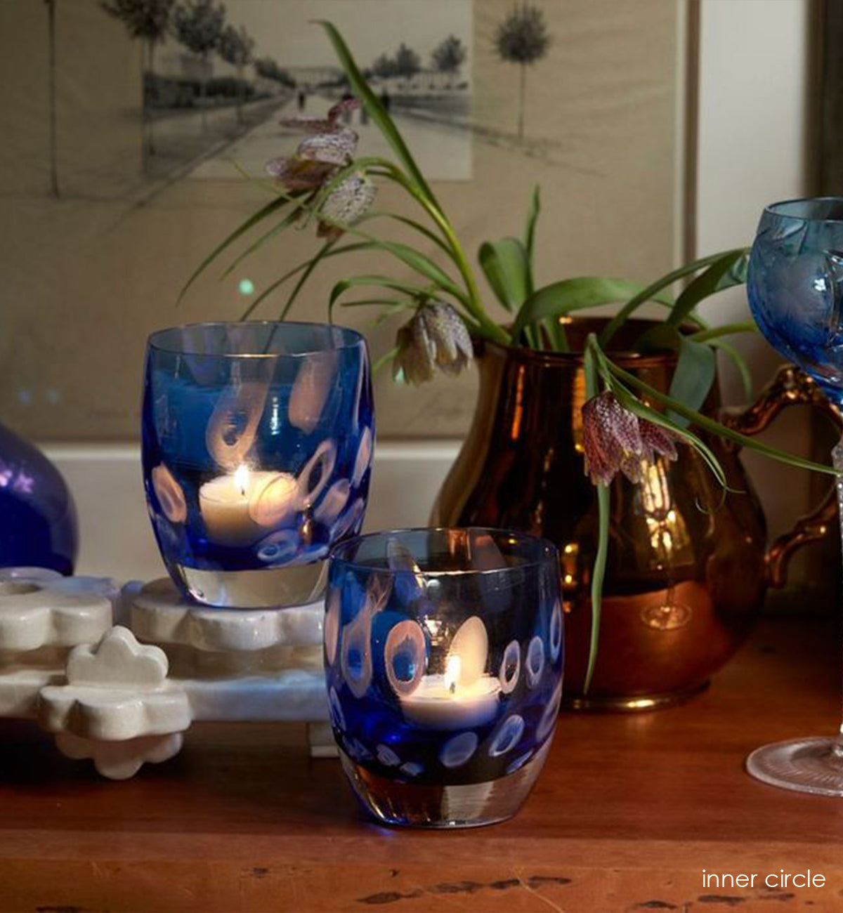 two inner circle, translucent dark blue and white murrini hand-blown glass votive candle holders on a wood dining room table with blue drinking glass, vase and flowers.